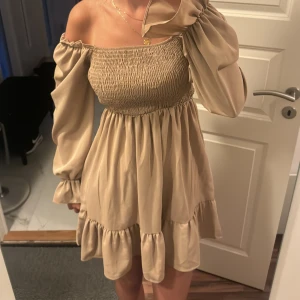 Sommarklänning  - beige  - Supersöt beige kortklänning med smockad överdel och volang nertill. Klänningen har långa ärmar med resår vid ärmslut och fyrkantig ringning. Midjan är stretchig 