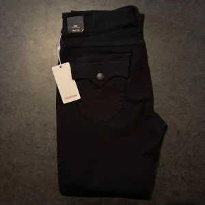 Svarta True Religion Ricky jeans - Svarta jeans från True Religion, modell Ricky. Klassisk femficksdesign med snygga detaljer på bakfickorna och logopatch bak. Jeansen har rak passform och är tillverkade i ett mjukt bomullstyg med lite stretch för extra komfort. Helt oanvända med prislapp. ställ frågor om du har! :D pris kan diskuteras.