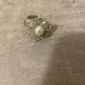 Ring med pärla och strass - Unik silverfärgad ring med stor vit pärla i mitten och glittrande strass i ett blommigt mönster runt om. Ringen har en bred, chunky design som verkligen sticker ut och ger en lyxig känsla. Perfekt för dig som vill ha något extra till din stil.