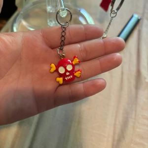 Röd dödskalle nyckelring med hjärtben - Nyckelring med en röd dödskalle och korsade ben formade som gula hjärtan. Dödskallen har vita ögon och tänder, och hänger i en metallkedja. Perfekt accessoar för dig som gillar unika detaljer.