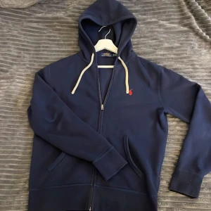 Polo Ralph lauren hoodie - Snygg marinblå hoodie från Polo Ralph Lauren med dragkedja och vit snörning i huvan. Klassisk röd logga broderad på bröstet. Perfekt för dig som vill ha en lite sjystare hoodie!