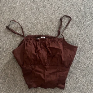 Brun linnetopp från H&M Divided - Säljer en brun linnetopp från H&M Divided i storlek 32. Toppen har tunna axelband och smockad resår upptill för en skön passform. Perfekt till sommaren och enkel att matcha med jeans eller kjol.