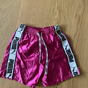 Rosa Puma shorts med logoband - Snygga glansiga rosa shorts från Puma med vita logoband längs sidorna och matchande snörning i midjan. Shortsen har elastisk midja och är fodrade med mesh för extra komfort. Perfekta för sommarens alla aktiviteter.