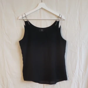 Svart linnetopp med kedjeband AX Paris - Stilren svart linnetopp från AX Paris med tunna kedjeband som axelband. Toppen har en rak passform och är tillverkad i ett lätt, flowy material som ger en snygg siluett. Perfekt att matcha med jeans eller kjol för en cool look.
