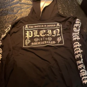 Svart Plein hoodie med tryck - Svart hoodie från Philipp Plein med stora vita tryck på bröstet och ärmarna. Tröjan har huva med ljus insida och metallmärke på huvan. Snygga detaljer med gotisk text och logga på ryggen. Perfekt för dig som gillar streetwear och statement-plagg.