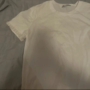 Vit t-shirt med pärldetaljer H&M - Vit t-shirt från Zara med korta ärmar och snygga pärldetaljer längst ut på ärmsluten. Klassisk rund halsringning och rak passform. Perfekt för dig som gillar enkla plagg med en twist. Tillverkad i mjuk bomull för skön känsla.