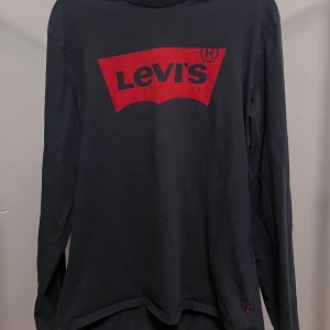 Svart långärmad Levi's tröja - Svart långärmad tröja från Levi's med det klassiska röda logotrycket framtill. Tröjan är i mjuk bomull och har en avslappnad passform. Perfekt för dig som gillar en enkel men ikonisk stil med tydlig branding.