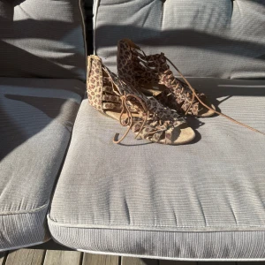 Leopardmönstrade sandaler från Mywear - Snygga sandaler från Mywear i leopardmönstrad mocka-look. Skorna har snörning framtill, öppen tå och platt sula. Perfekta för dig som vill sticka ut med ett djurmönster och gillar detaljer som ger extra edge till outfiten.