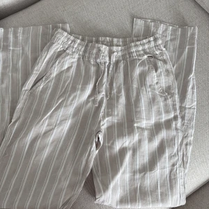 Randiga linnebyxor från 157, beige - Snygga beige linnebyxor från 157 med vita vertikala ränder. Byxorna har elastisk midja med snörning, raka ben och sidofickor samt en bakficka. Perfekta för en chill och luftig stil under varma dagar.