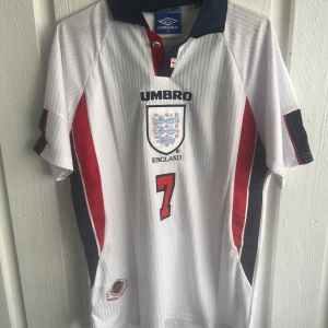 England Beckham 7 fotbollströja Umbro - England fotbollströja från Umbro med Beckham och nummer 7 på ryggen. Vit med tunna vertikala ränder, röda och mörkblå detaljer längs sidorna och ärmarna. Klassisk krage i mörkblått. Broderat England-emblem på bröstet. Perfekt för dig som älskar fotboll och retrostil.