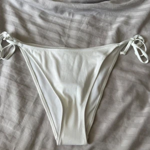 Vit bikiniunderdel med knyt från H&M - Snygg vit bikiniunderdel från H&M i modellen tie tanga. Klassisk tanga-form med smala knytband i sidorna som gör att du kan justera passformen. Tillverkad i mjukt och stretchigt material som passar perfekt till stranden eller poolen.