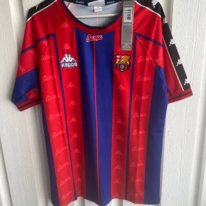 Barcelona Ronaldo 9 Kappa tröja - Klassisk FC Barcelona fotbollströja från Kappa med Ronaldo 9 på ryggen. Tröjan har röda och blå vertikala ränder, klubbmärke på bröstet och Kappa-loggor på axlarna. Materialet är lätt och andas, perfekt för match eller samling. Snygg retrostil med korta ärmar.