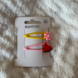 Färgglada hårspännen från Montini - Två söta hårspännen från Montini, ett gult med en rosa blomma och ett rosa med en vattenmelon. Perfekta för att ge din frisyr en lekfull och färgglad touch. Säljs på originalkort från Ur & Penn.