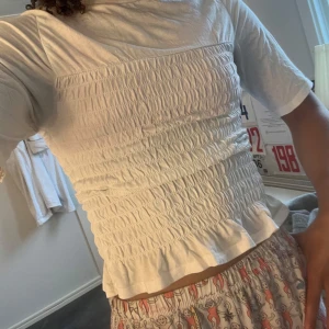 Vit smockad topp från H&M Divided XS - Supersöt vit topp från H&M Divided i storlek XS. Toppen har smockat liv, volangkant nertill och halvlånga ärmar. Gjord i mjuk bomull, perfekt till jeans eller kjol för en chill look.