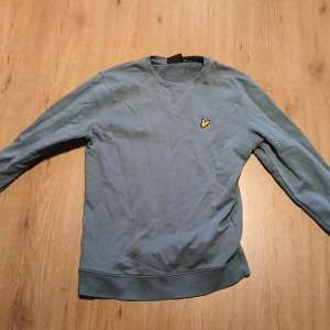 Blå tröja från Lyle & Scott - Snygg blå sweatshirt från Lyle & Scott med klassisk rund hals och gul logga på bröstet. Tröjan har långa ärmar och ribbade muddar vid ärmslut och nederkant. Perfekt för en chill och stilren look. Pris kan diskuteras 