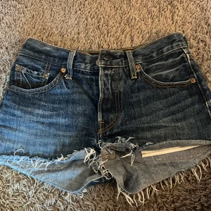 Levi's jeansshorts - Snygga blå jeansshorts från Levi's med fransig rå kant och klassiska fem fickor. Shortsen är mid rise, knäppning med knappar och slitna detaljer framtill. Perfekta för sommaren och ger en cool vintagekänsla.