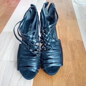 Svarta klackade sandaler i skinn - Svarta sandaler i äkta skinn med höga klackar och öppen tå. Snyggt flätade remmar framtill och dragkedja bak för enkel påtagning. Guldigt emblem på sulan ger en exklusiv touch. Perfekta för dig som vill sticka ut.