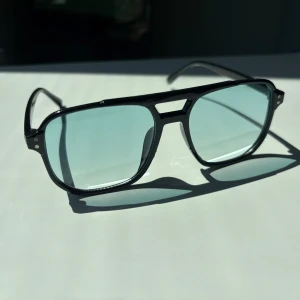 Svarta pilotbågar med blåtonade glas - Snygga pilotinspirerade glasögon med svart ram och ljusblåtonade glas. Bågarna har en cool dubbelskena över näsan och markerade kanter. Perfekta för dig som vill sticka ut med en retrovibe.