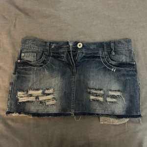 Lågmidjad jeanskjol  - Säljer en kort jeanskjol i mörkblå denim med rå kant och slitna detaljer framtill och på bakfickorna. Kjolen har klassiska fickor, bälteshällor och dragkedja med knapp. Perfekt för dig som gillar en edgy och avslappnad stil.