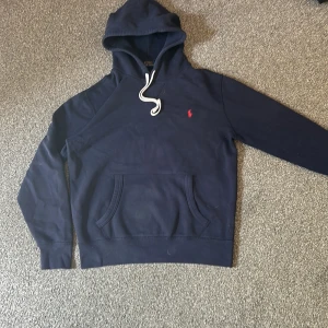 Mörkblå hoodie från Polo Ralph Lauren - Säljer en mörkblå hoodie från Polo Ralph Lauren med vit snörning i huvan och klassisk röd logga broderad på bröstet. Tröjan har känguruficka framtill och är tillverkad i mjuk bomull. Perfekt för en chill och stilren look.