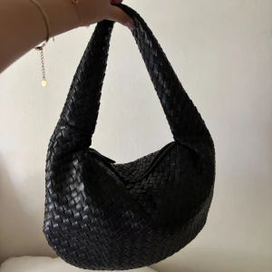 Svart flätad axelväska i skinn - Säljer en svart axelväska med snygg flätad design och bred axelrem. Väskan har dragkedja upptill. Perfekt storlek för att få plats med allt du behöver och ger en cool vibe till din outfit.