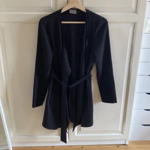 Svart kofta från Vero Moda med bälte - Stilren svart kofta från Vero Moda med lång ärm och knytbälte i midjan. Koftan har öppen front och två fickor på sidorna. Storlek 36. I fint skick, har knappt använt den. 