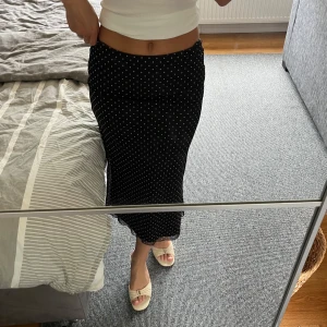 Svart prickig långkjol från BikBok - Svart långkjol från BikBok med vita prickar och lätt volangkant nertill. Kjolen har en rak passform och är gjord i ett tunt, flowy material som känns skönt mot huden. Perfekt för dig som gillar en klassisk men ändå trendig look.