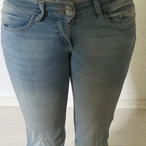  jeans - Säljer ett par ljusblå jeans med bootcut och slitna detaljer vid knäna. Jeansen har hög midja, dubbla knappar framtill och bakfickor med lock och knapp. Perfekta för en avslappnad och trendig look.
