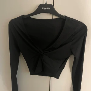 Svart croppad topp med knyt från KappAhl - Svart långärmad croppad topp med snygg v-ringning och knyt framtill. Toppens ribbade material ger en cool struktur och den sitter tajt och smickrande. Perfekt för dig som gillar trendiga och stilrena plagg.