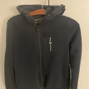 Svart Sail Racing hoodiejacka 176 - Svart hoodiejacka från Sail Racing med dragkedja framtill och bröstficka med logga. Jackan har huva med snörning och är tillverkad i mjukt material, perfekt för chill dagar. Snygg och enkel design som funkar till det mesta.