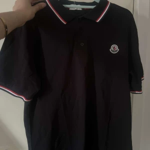 Svart Moncler pikétröja med röd/vit kant - Svart pikétröja från Moncler med klassisk krage och knäppning framtill. Tröjan har röda och vita ränder på krage och ärmslut samt Moncler-logga på bröstet. Tillverkad i bomull och har korta ärmar. Perfekt för en clean och sportig look.