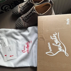 Christian Louboutin skor - Unika grå sneakers från Christian Louboutin i mocka med ikoniska röda sulor och coola svarta nitar på tån. Skorna har snörning och rund tå, samt exklusiv känsla med lyxiga material och detaljer. Perfekta för dig som vill sticka ut med din stil. Skriv vid minsta fundering! Storleken passar 44-46