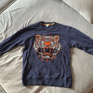 Mörkblå Kenzo tröja med tigertryck  - Säljer en mörkblå crewneck från Kenzo i bomull med ikonisk tigerbrodyr framtill i orange, vitt och blått. Tröjan har ribbade muddar och rund hals. Perfekt statement piece för dig som vill sticka ut med stil.
