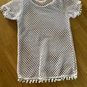 Vit nätblus med bollar, stl 140 - Superfin vit nätblus i mesh med korta ärmar och söta bollar längs ärmslut och nederkant. Perfekt för stranden som en strand klänning för barn. Tillverkad i bomull och elastan för extra stretch och komfort.