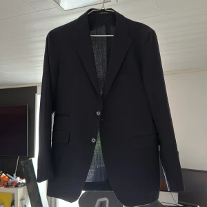 Kavaj från Tagliatore - Snygg svart kavaj från Tagliatore, tillverkad i Italien. Klassisk modell med två knappar framtill, diskreta fickor med lock och elegant snitt. Tunt fodrad insida och stilrena detaljer. Perfekt för dig som vill ha en tidlös och stilren look. Liten defekt på armar som knappast syns och gå att laga på sista bilden. Köpt för 7499!