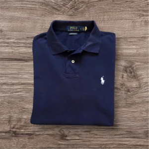Marinblå pikétröja från Ralph Lauren - Klassisk marinblå pikétröja från Ralph Lauren med vit broderad logga på bröstet. Tröjan har krage, två knappar och är tillverkad i mjuk bomull. Perfekt för en stilren och avslappnad look.