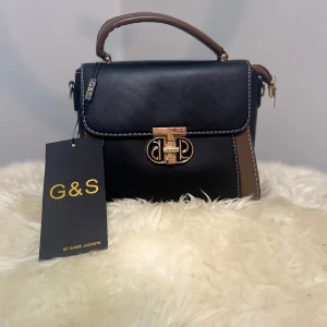 Svart och brun handväska från G&S - Snygg handväska från G&S i svart och brunt skinn med guldfärgat spänne framtill. Väskan har ett stadigt handtag och medföljande axelrem. Kontrastsömmar och stilren design gör den perfekt för dig som gillar klassiska accessoarer.