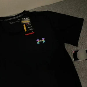 Svart t-shirt från Under Armour - Svart t-shirt från Under Armour med klassisk passform och korta ärmar. Materialet är lätt och andas, perfekt för träning. Framtill finns en snygg, reflekterande logga i regnbågsfärger som ger en sportig vibe.