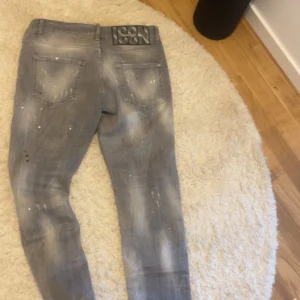Grå Dsquared2 ICON jeans - Säljer ett par grå Dsquared2 ICON jeans med coolt sliten tvätt och vita färgstänk. Byxorna har raka ben, klassiska bakfickor och stor svart patch med 'ICON' på linningen. Perfekta för dig som vill sticka ut med en streetig vibe.