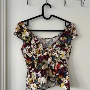 Blommig omlottopp från Guess XS - Supersöt omlottopp från Guess i storlek XS med färgglatt blommönster. Toppen har korta ärmar, v-ringning och en figurnära passform. Perfekt för dig som gillar romantiska vibes och vill sticka ut med en unik topp.☀️💗🥂