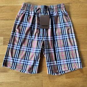 Rutig Burberry shorts med snörning - Snygga shorts från Burberry med klassiskt rutigt mönster i beige, svart, vit och rosa. De har elastisk midja med svart snörning för perfekt passform. Perfekta för sommaren och ger en lyxig vibe till din outfit.