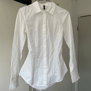 Vit skjorta med öppen rygg och knyt - Säljer en vit skjorta  med lång ärm, klassisk krage och knappar framtill. Baksidan har en stor öppen rygg och knytband i midjan för en unik look. Skjortan är figursydd och tillverkad i bomull, perfekt för dig som vill sticka ut.
