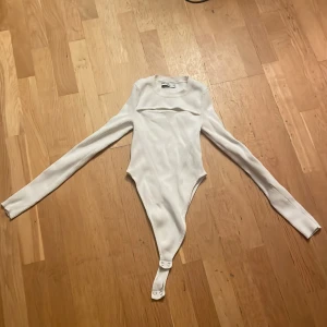 Vit ribbad bodysuit från Sister Knitwear - Supersnygg vit ribbad bodysuit från Sister Knitwear med lång ärm och rund hals. Bodyn har en cool detalj över bröstet och knäppning nertill. Perfekt att styla med jeans eller kjol för en trendig look.