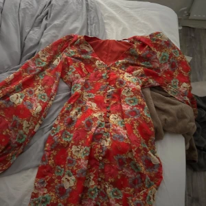 Röd blommig kortklänning med v-ringning - Superfin röd kortklänning med blommigt mönster och v-ringad hals. Klänningen har långa, puffiga ärmar och en figurnära passform. Från adoore med ett mönster och färg som inte längre sälj 🥰