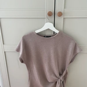 Beige stickad topp med knyt från Zara - Superfin beige stickad topp från Zara med korta ärmar och rund hals. Toppen har en snygg knytdetalj i sidan som ger en cool och modern look. Perfekt att styla med jeans eller kjol för en avslappnad vibe.