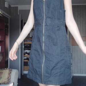 Svart ärmlös klänning med dragkedja - Svart ärmlös kortklänning med grov dragkedja framtill och coola fickdetaljer på sidorna. Klänningen har en enkel, rak passform och är tillverkad i ett stadigt bomullstyg. Perfekt för dig som gillar en stilren och edgy look.