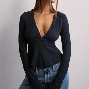 svart omlottopp med v-ringning - Snygg svart omlottopp med djup v-ringning och långa ärmar. Toppen har ribbad struktur och knytning i ryggen för en extra detalj. Perfekt att styla med jeans för en cool look.