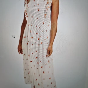 Vit långklänning med broderade blommor - Superfin vit långklänning med broderade orange och röda blommor. Klänningen har volangdetaljer vid axlarna, knappar framtill och är ärmlös. Perfekt för sommarens alla tillfällen, med ett luftigt och romantiskt uttryck. Materialet känns mjukt och svalt.