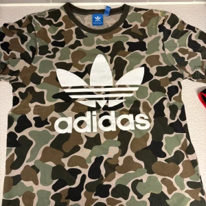 Adidas camo t-shirt med logga - Snygg t-shirt från Adidas med klassisk vit trefoil-logga framtill. T-shirten har ett fett camouflage-mönster i grönt, brunt och svart. Rund halsringning och korta ärmar. Perfekt för dig som vill sticka ut med streetstyle. Det är dock några små klorinfläckar på 