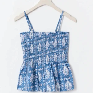 Blå blommig linnetopp med volang - Supersöt blå linnetopp med vita och ljusrosa blommor. Smockad upptill och volang nertill, tunna axelband och luftig känsla. Perfekt för varma dagar och ger en chill sommarvibe. Går att justera banden så de blir en tub topp 